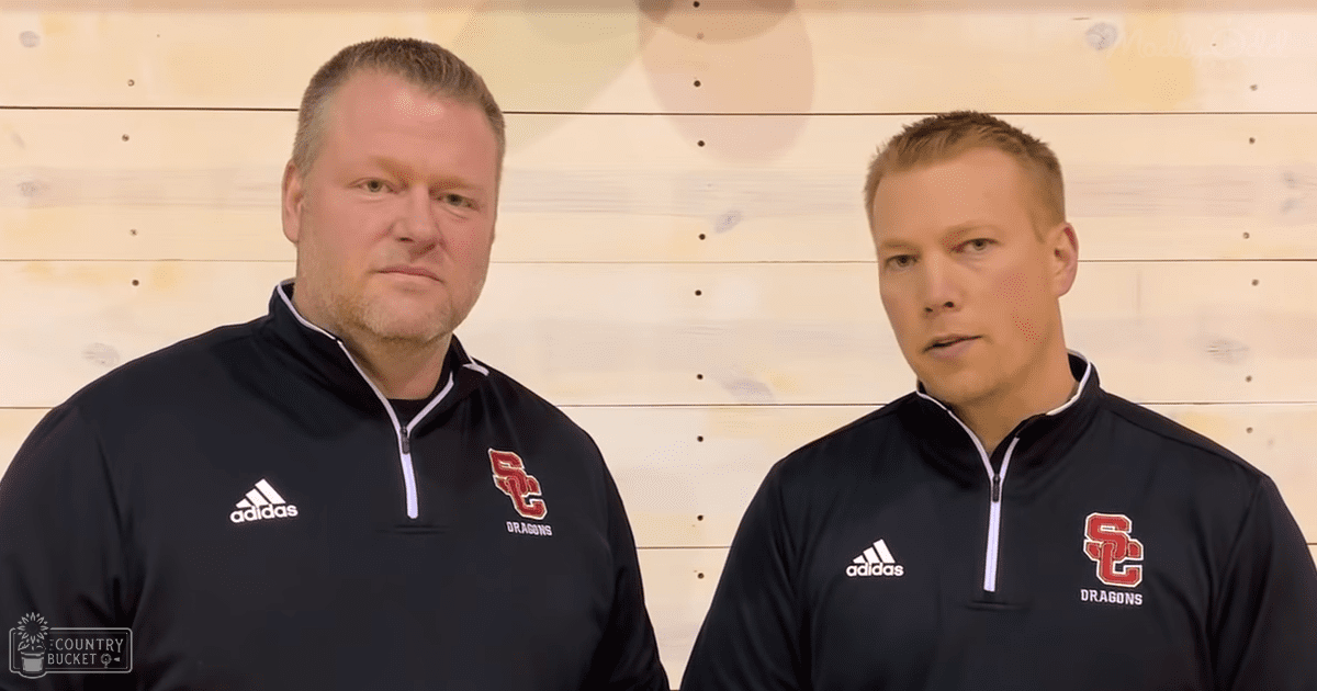 Two adults in black Adidas quarter-zip jackets with 'SC Dragons' logo, standing indoors with light-colored wooden planks as the backdrop.