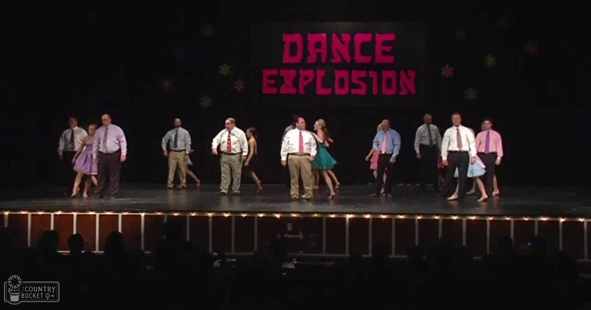 A group performance at "Dance Explosion" in an auditorium with performers dressed in various shades of blue, pink, and red, under stage lighting.