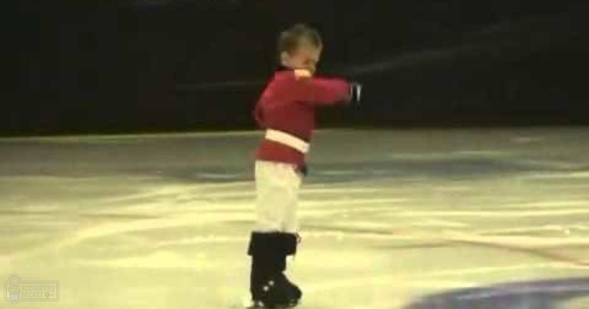 A toddler in a maroon sweater with white trim, white pants, and black roller skates is skating indoors on a smooth rink under artificial lighting.