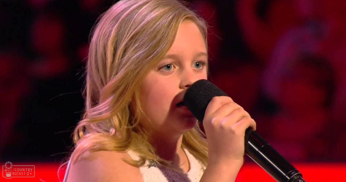 Young girl wearing a white sleeveless top, singing into a microphone during a talent show performance with bright stage lights.