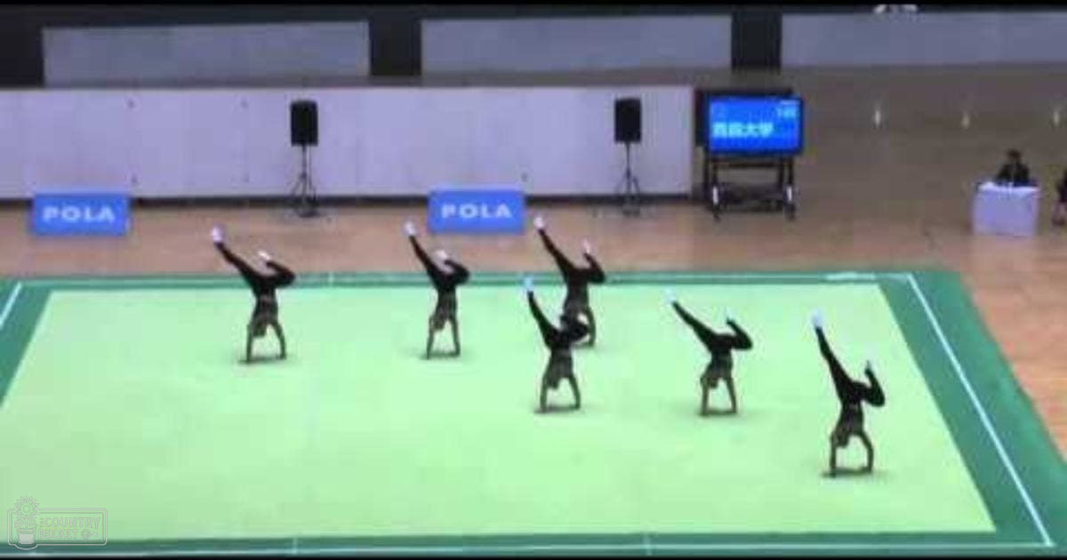 Young female gymnasts performing handstands in a brightly lit indoor gymnasium with blue sponsor signage.