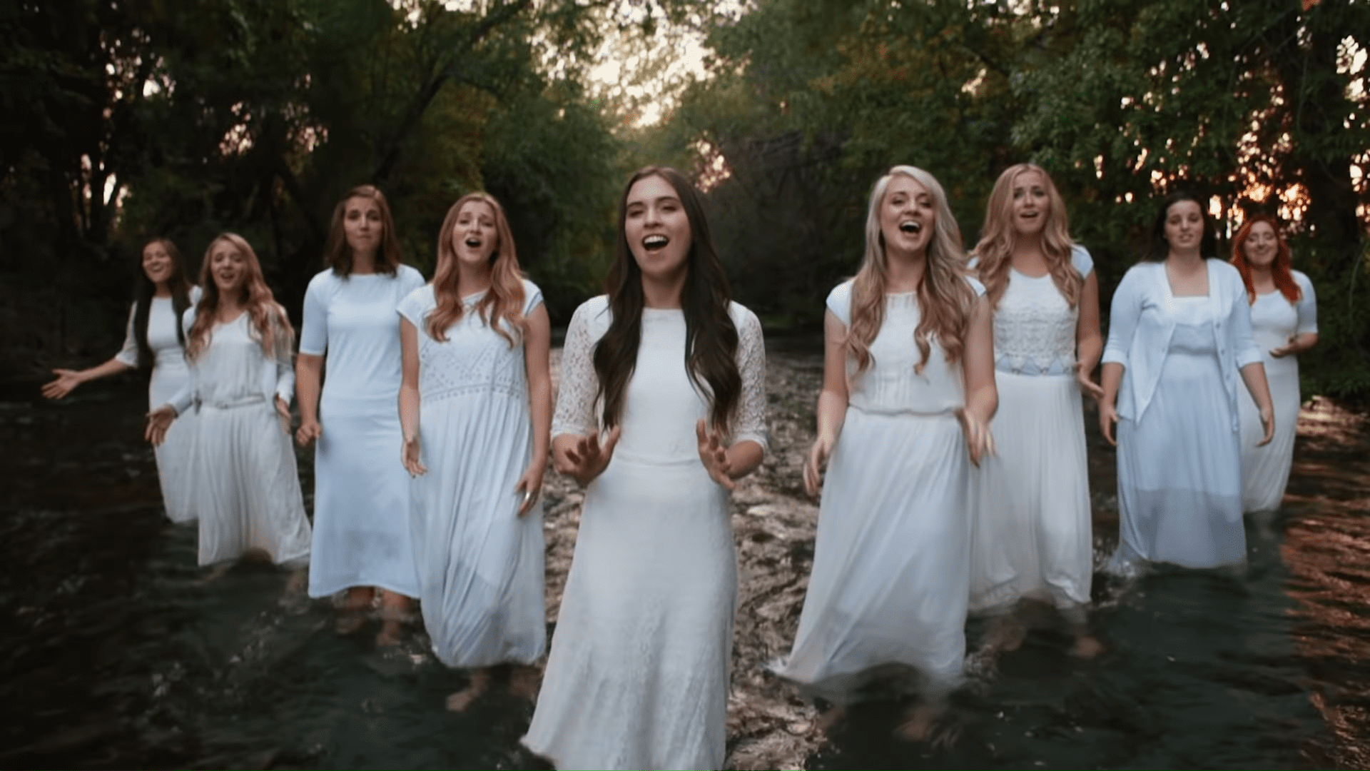 A group of young adult women singing in a natural outdoor setting with trees and water, dressed in white knee-length lace dresses.