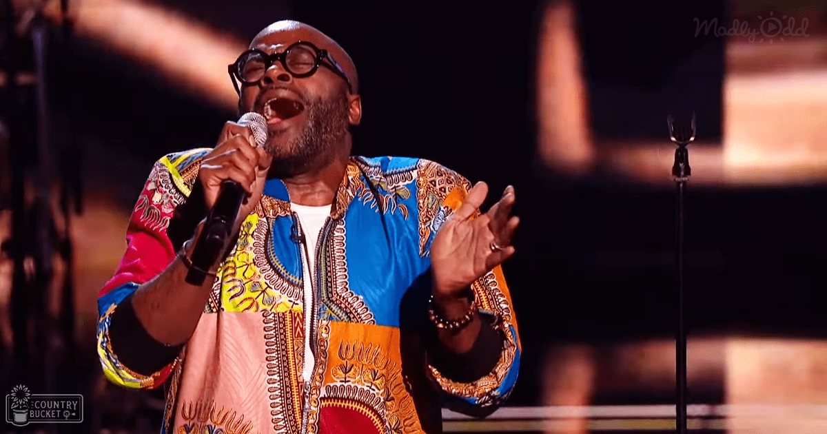 A middle-aged male singer performing on a dimly lit stage, wearing a colorful traditional African print garment with vibrant blue, yellow, orange, and red hues.