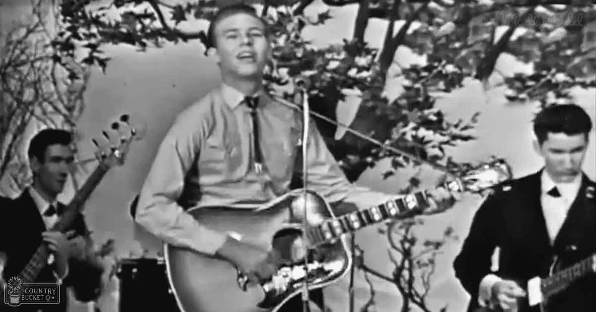 Young adult male singer playing guitar outdoors with trees in the background, accompanied by a bassist and another guitarist in formal attire.
