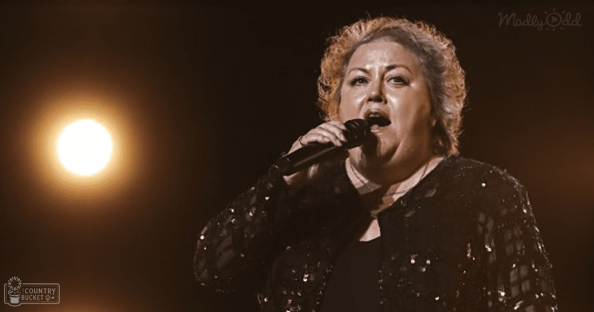 A middle-aged female singer wearing a black sequined jacket performs into a microphone at an indoor concert venue with focused stage lighting.