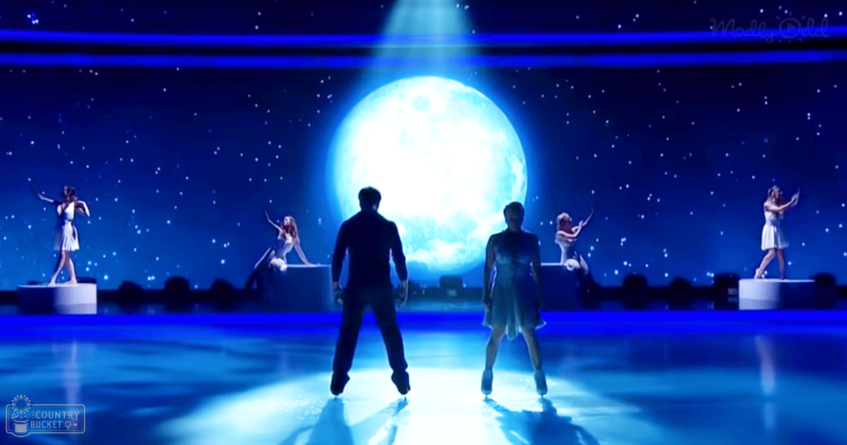 An ice skating performance at a nighttime indoor venue with figures in white dresses against a backdrop of illuminated moon and stars.