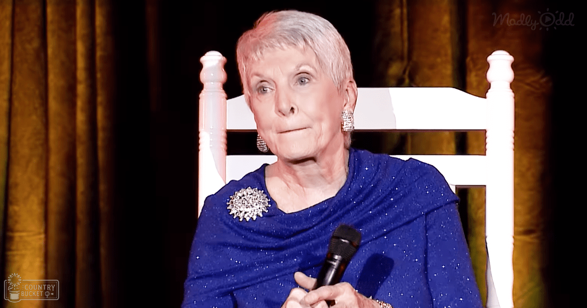 An older adult woman wearing a blue, glittery formal dress with long sleeves and a large silver brooch on her left shoulder stands indoors at what appears to be an event or gathering. A white ornate chair is visible behind her.