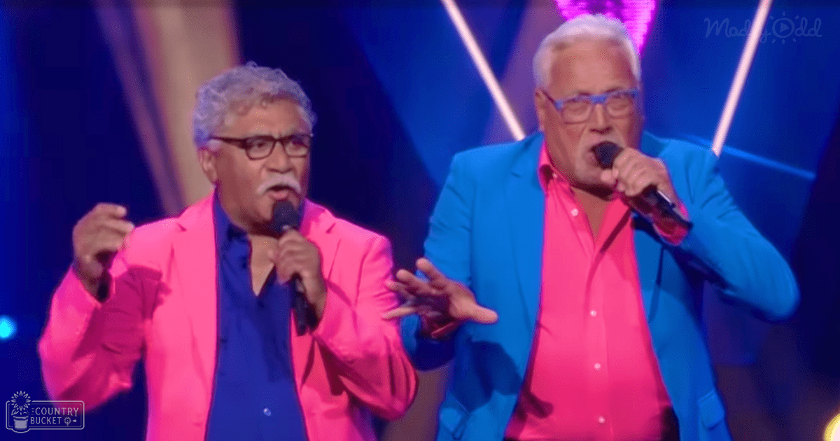 Two older adult male singers performing on a brightly lit stage at an indoor music event with vibrant blue and white lighting beams.