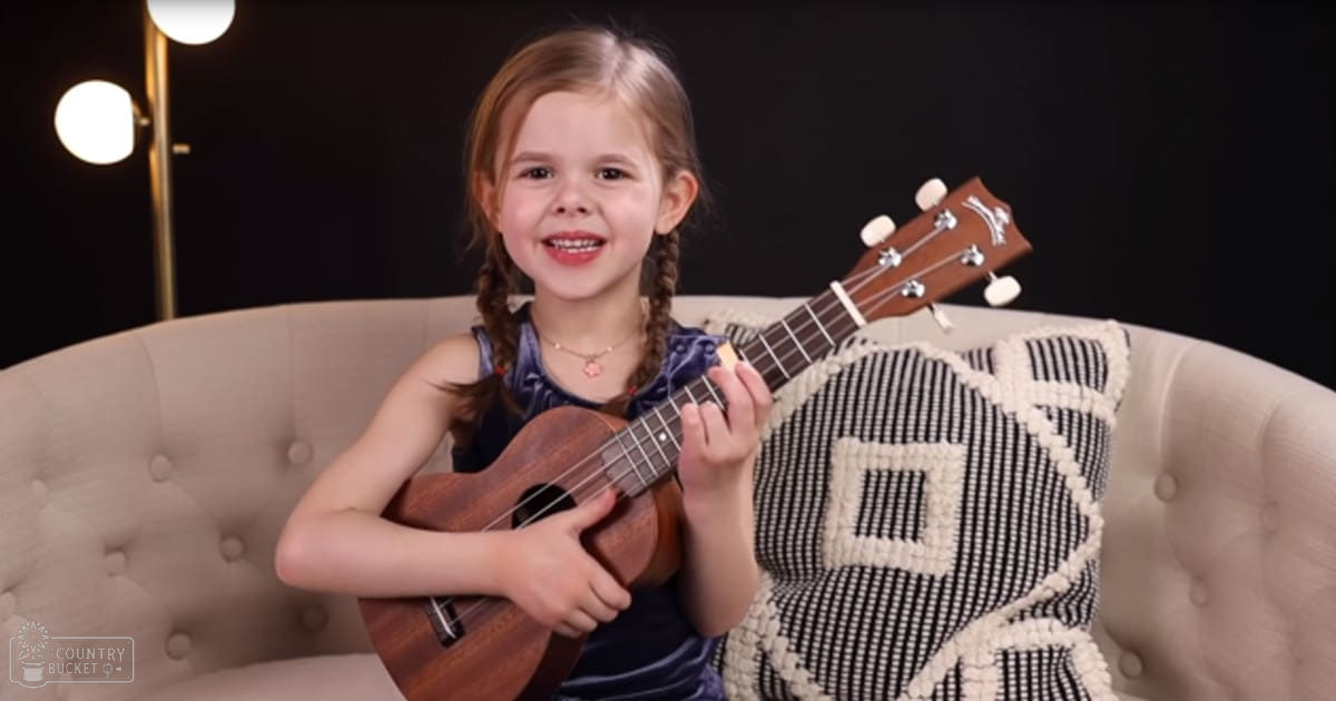 A toddler girl plays a ukulele in a cozy indoor living room with warm lighting, wearing a dark blue sleeveless top.