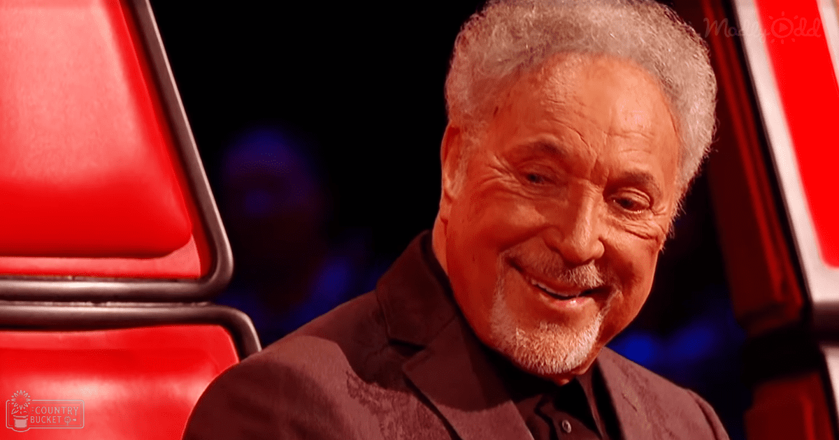 An older adult male in a dark blazer, black shirt, and red chair with metallic frame on a brightly lit stage setup.