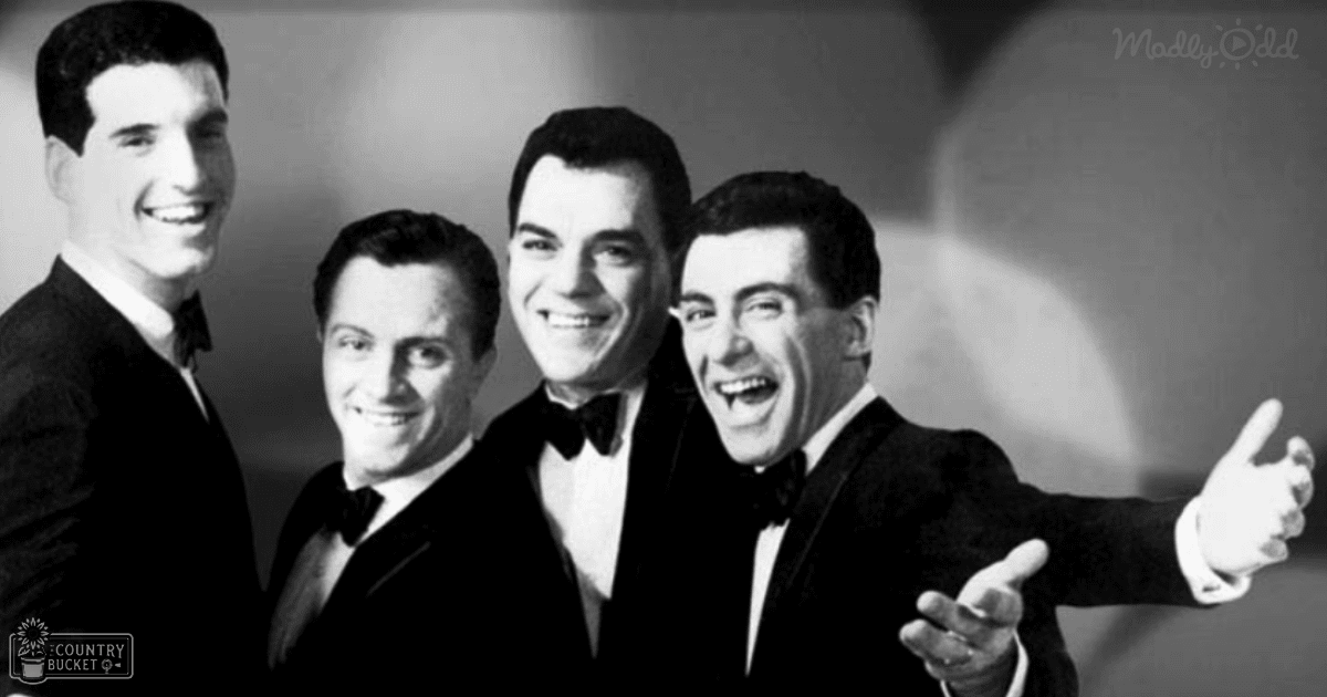 A group photo of four young adult males dressed formally in dark suits with white shirts and black bow ties, taken indoors under soft artificial light.