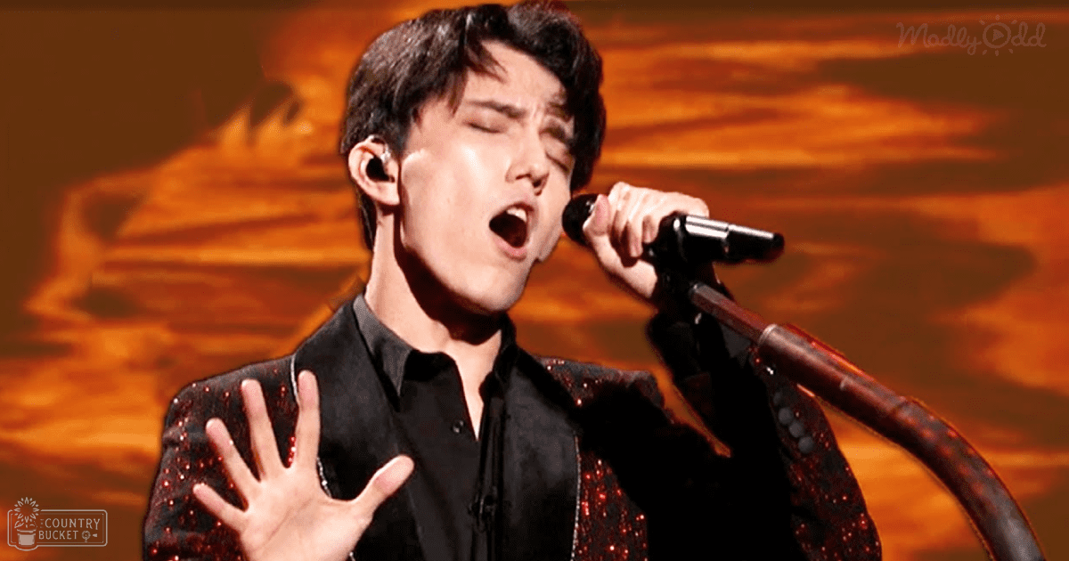 Young adult male in a button-down shirt with a sequined jacket performing on stage under warm, orange-toned lighting.