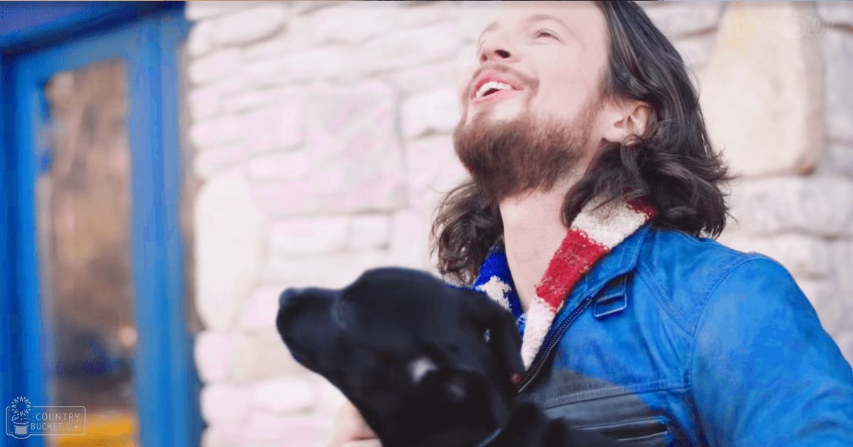 A male adult in a blue leather jacket with red and white patterned collar stands outdoors near a stone wall with a black Labrador Retriever. The setting suggests an autumn or spring day, with soft natural light.