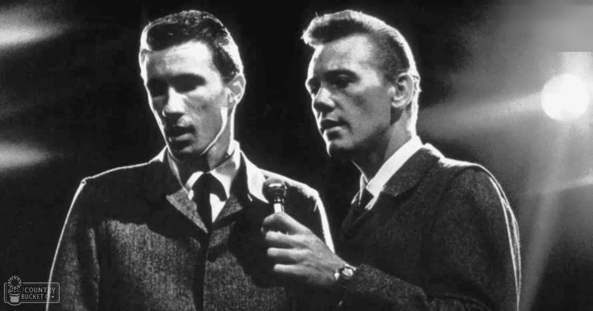 Adult male singer wearing a dark textured suit with tie and light shirt on stage with another adult male in a dark sweater and light shirt, holding a microphone under dim stage lighting.
