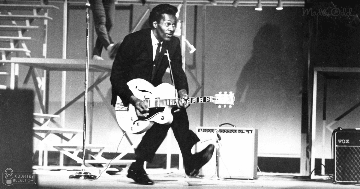 An adult male performer in a black suit jacket and trousers, playing an electric guitar on stage with a VOX amplifier and microphone stand, under evening or night lighting.