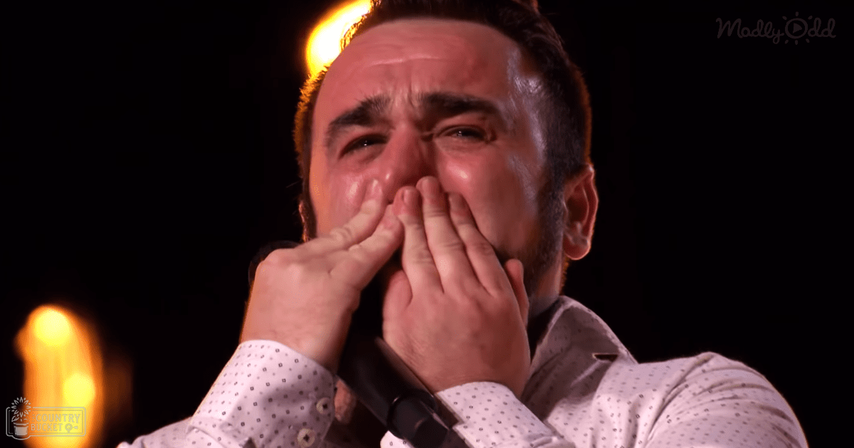 A middle-aged man wearing a white polka-dot shirt performs on stage under dim lighting with bright yellowish-orange stage lights.