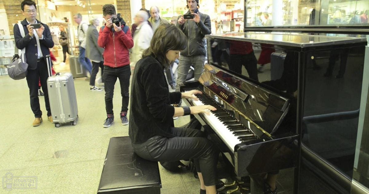 A woman playing a glossy black piano with gold accents, surrounded by people in an indoor public space with natural light and travel luggage.