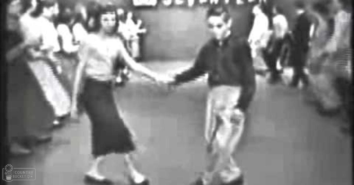 Young adults dressed in early 20th-century attire dance together indoors under artificial lighting at a social gathering.