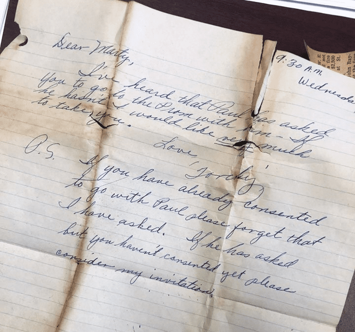 An indoor setting with a handwritten letter addressed to 'Marty' on a piece of lined paper, accompanied by a partially visible newspaper section showing text.