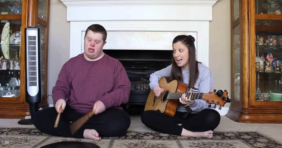 Young adult male singer in maroon sweatshirt, young adult female barefoot in gray shirt and black leggings, cozy living room setting with white fireplace and wooden cabinets.