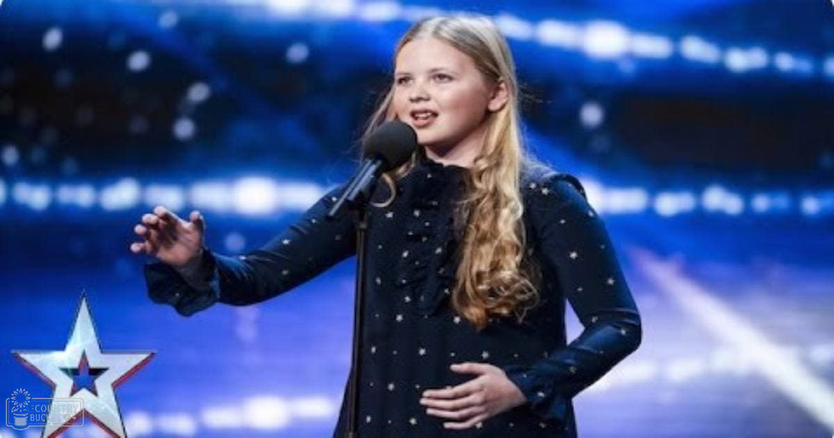 A young adult female with long blonde hair performs in a stage setting, wearing a dark blue dress with small white star patterns and ruffled details.