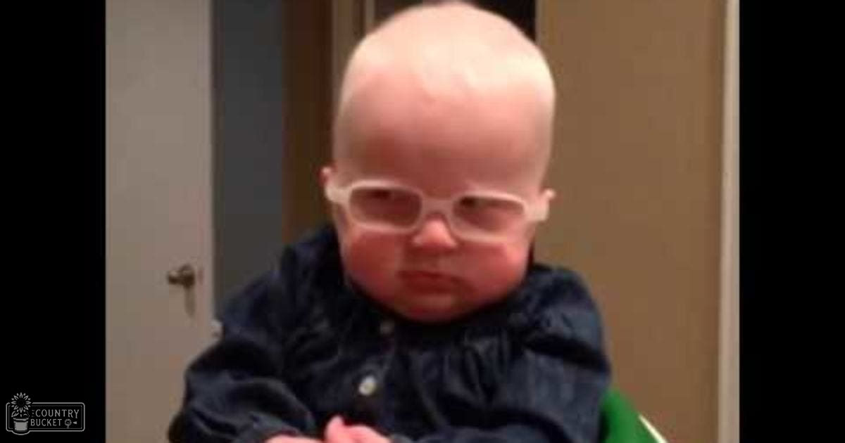 An infant wearing a dark blue button-up shirt or onesie, indoors with neutral-colored walls and a partially visible door.