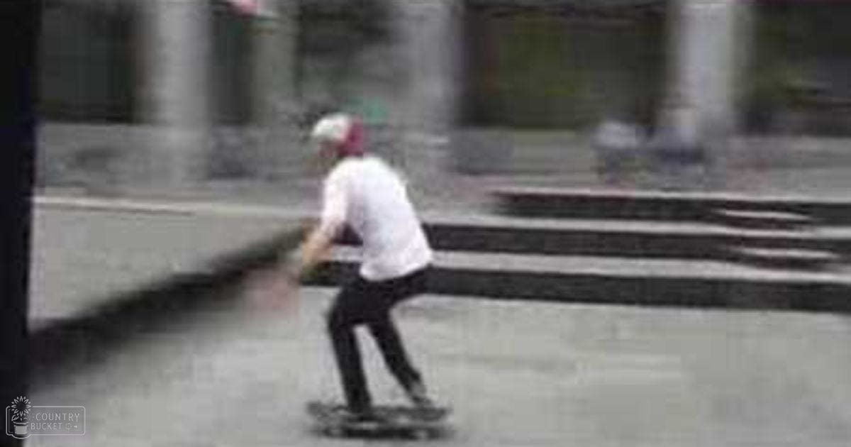 A young adult male skateboarder in an urban setting with stone steps and columns, wearing a white t-shirt, black pants, white sneakers, and a red, green, and white cap. The scene suggests daytime lighting.