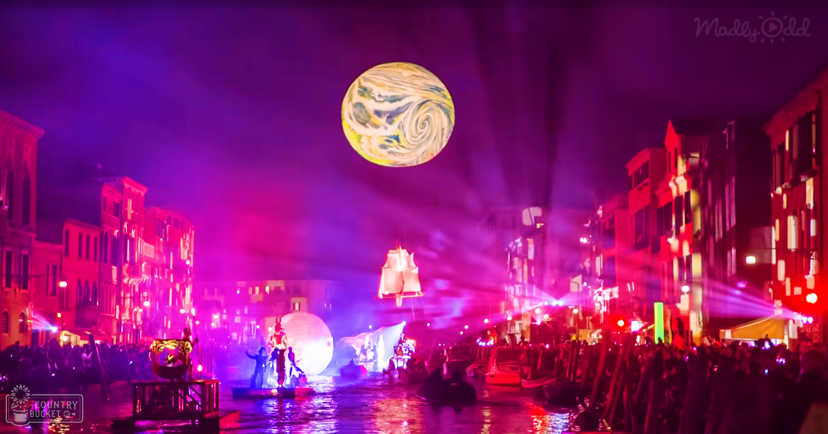 A vibrant nighttime festival scene in a canal city, likely Venice with colorful spotlights and illuminated structures along the waterway.