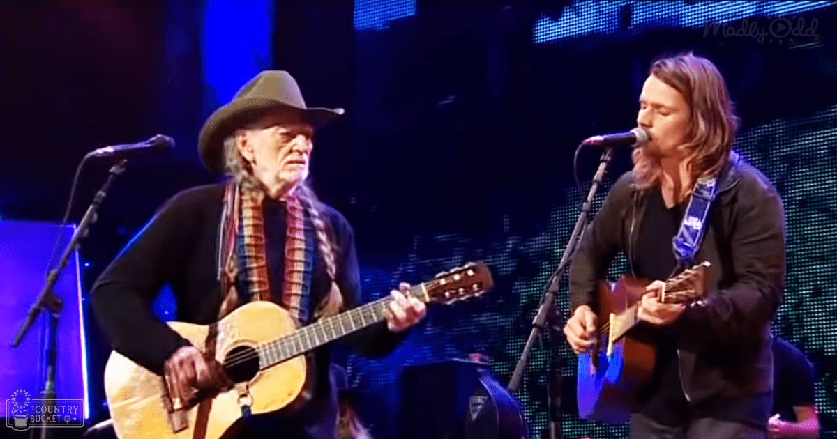 Musical performance scene at an indoor concert venue with older adult male in a wide-brimmed hat and patterned scarf, younger adult male in black jacket over dark shirt, digital screen with blue lighting, stage lights, acoustic guitars, blue tones dominating the setting.