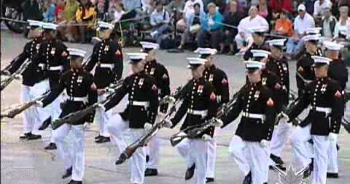 Military parade with uniformed men carrying rifles in an urban outdoor setting with spectators on a sunny day.