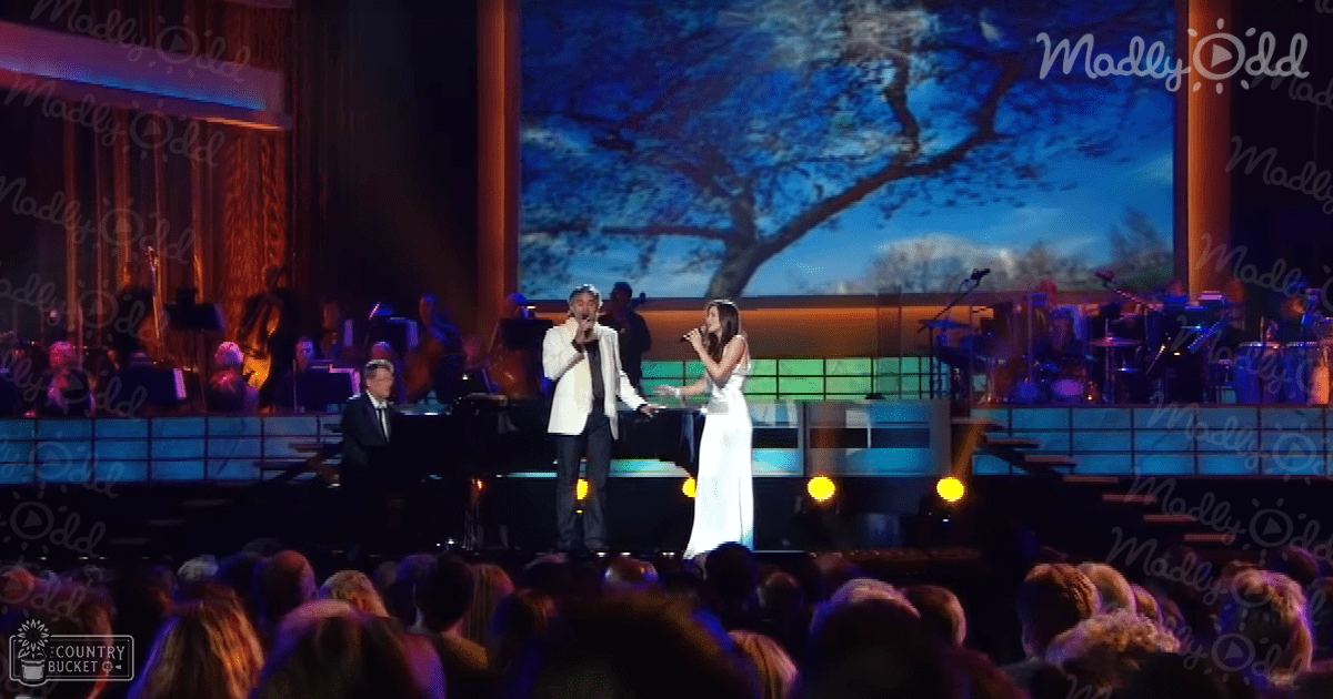 A male adult singer in a white blazer performs alongside a female adult in a long white dress, both holding microphones on stage with a grand piano and string players visible behind them. The background screen displays an outdoor scene.