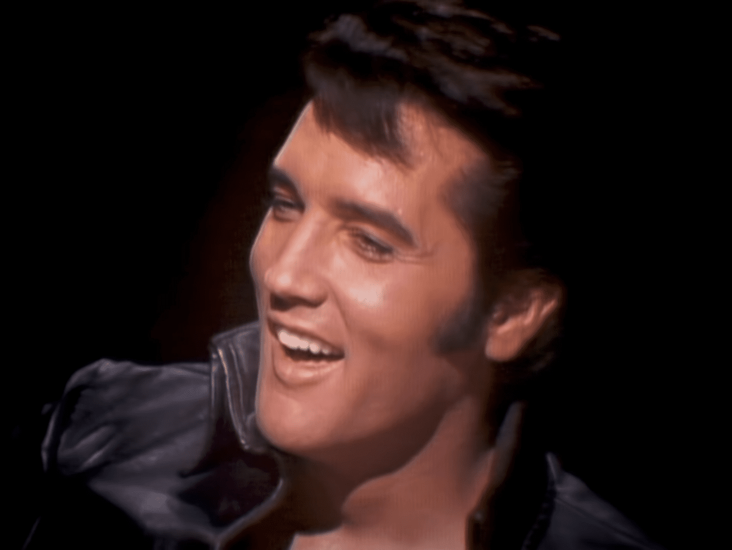 An adult male performer wearing a black leather jacket with puffy shoulders, standing indoors under dramatic stage lighting.