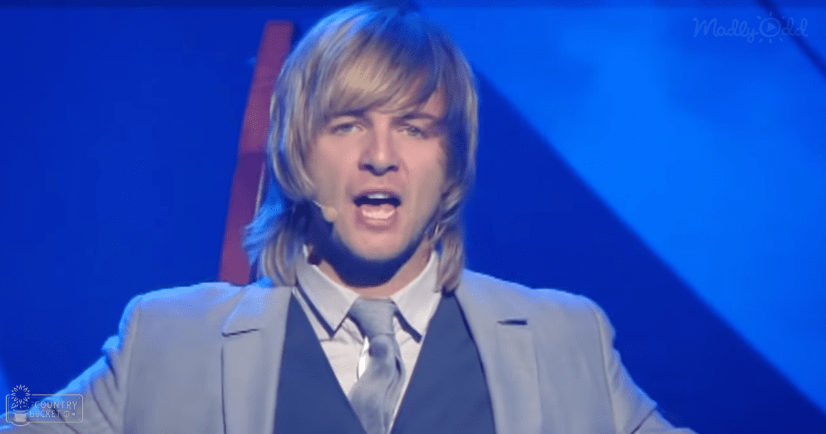 A young adult male wearing a light gray suit jacket on stage with a blue background, performing indoors under artificial lighting.