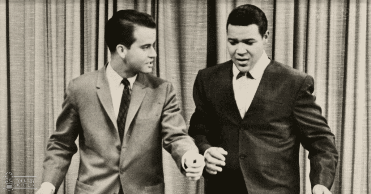 Two men in formal suits stand side by side; the setting appears to be an indoor studio or stage with a striped curtain backdrop, bright even lighting suggesting daytime.