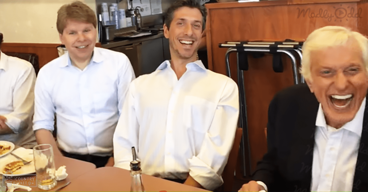 A group of adults in a casual dining setting, wearing light blue button-up shirts with crisp cotton fabric, white relaxed fit button-ups, and dark suit jackets over light blue dress shirts. Wooden table with plates of pancakes, coffee machine, and kitchen appliances visible.