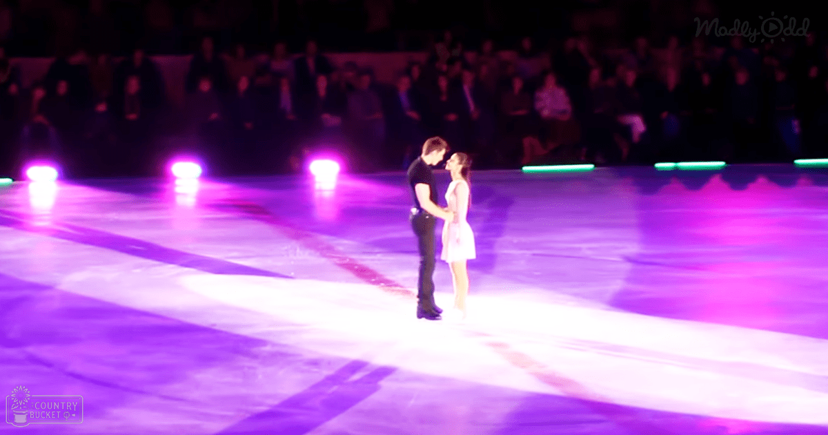 A couple performing on an indoor ice rink at a skating event or show; male in black shirt, female in light pink dress with spectators watching under artificial evening lighting.