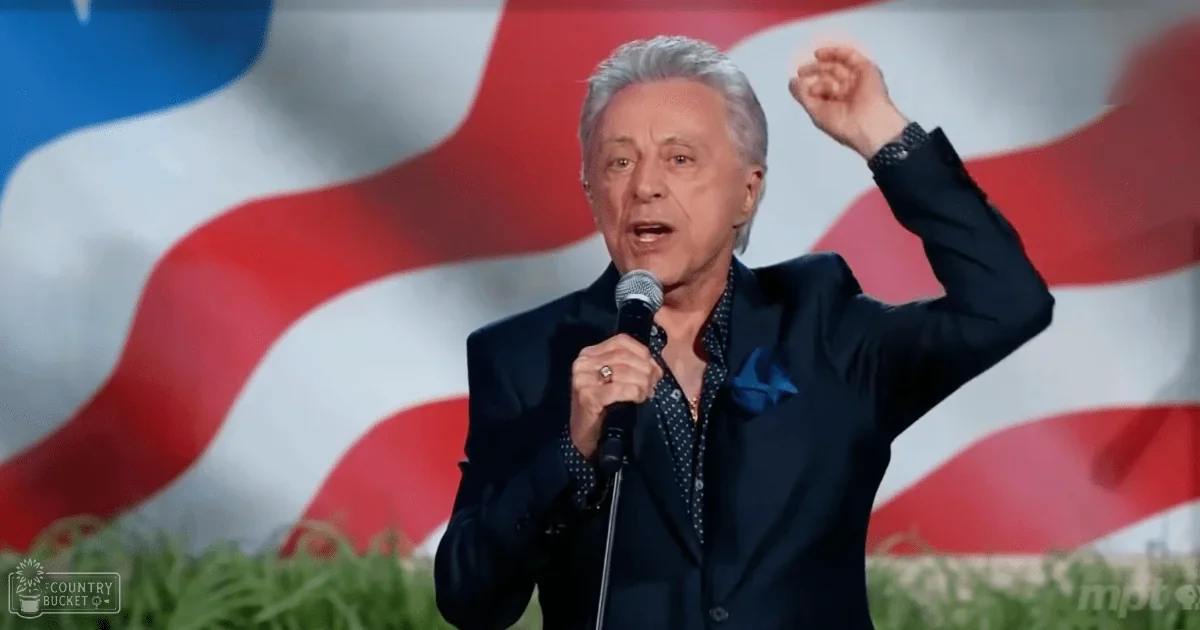 A middle-aged man in a dark blue suit with a polka dot shirt and blue pocket square stands before an American flag backdrop during what appears to be a public event.