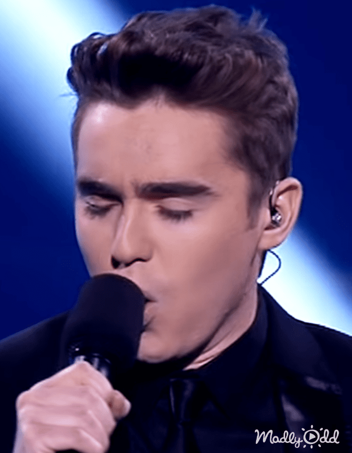 Young adult male in a dark-colored shirt with a bow tie on stage; 'Madly Odd' text visible.