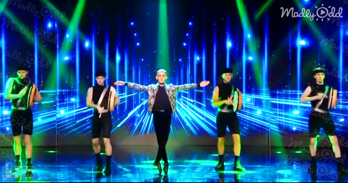 A young adult male performer in a patterned jacket with four background performers, all dressed casually on an indoor stage under vibrant blue and green lights at nighttime.