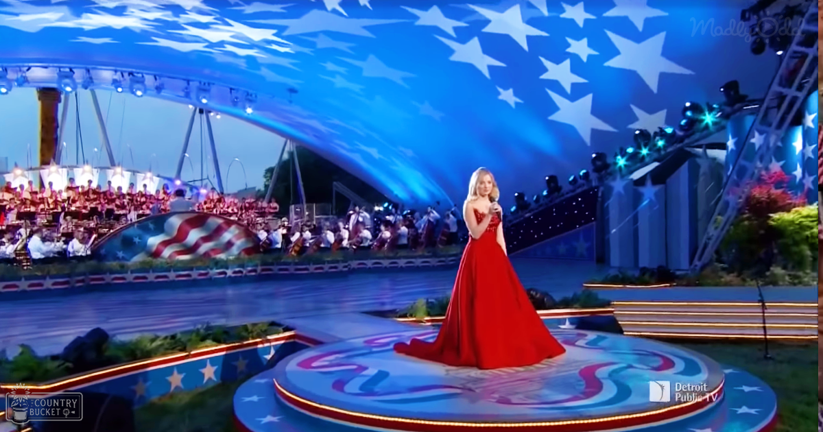An outdoor concert setting with an adult female in a flowing red gown at night, illuminated by stage lights with American flags and patriotic decorations.