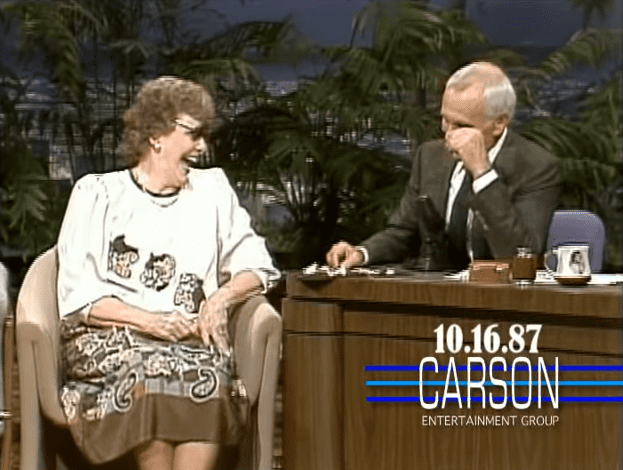 Talk show host interviewing an older adult guest in a studio setting with palm trees and evening lighting, suggesting a tropical or warm climate location.