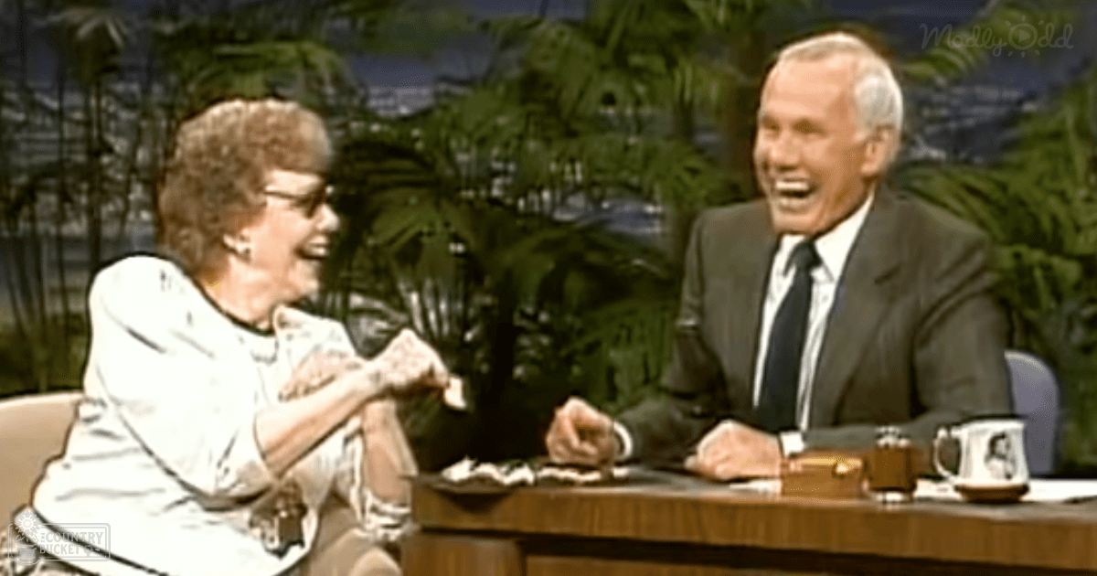 A lush green indoor studio with a talk show set, with two older adults in formal attire against bright and even artificial lighting.