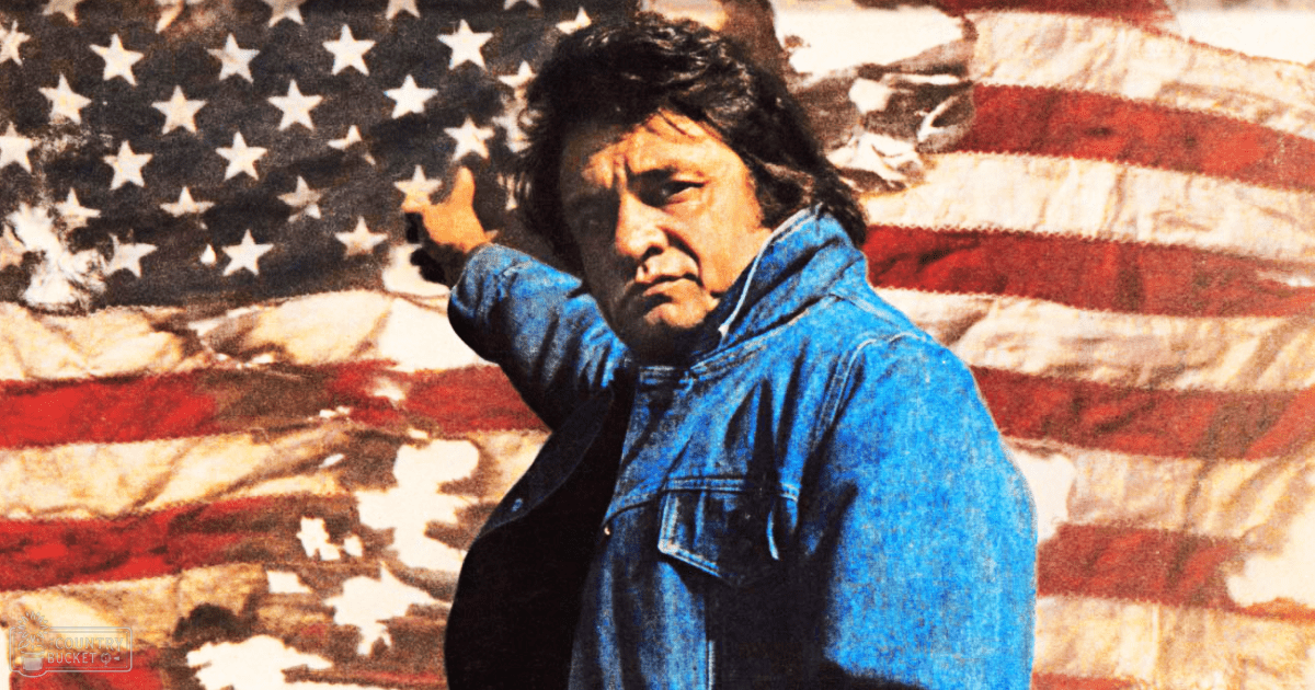 An adult male in a relaxed blue denim jacket with an American flag in the background, outdoors during daylight hours.