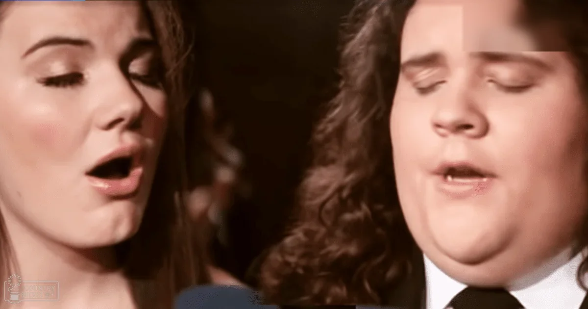 Indoor performance scene with young adult female in white top and middle-aged male in black tie against warm-toned background, possibly for music recording.