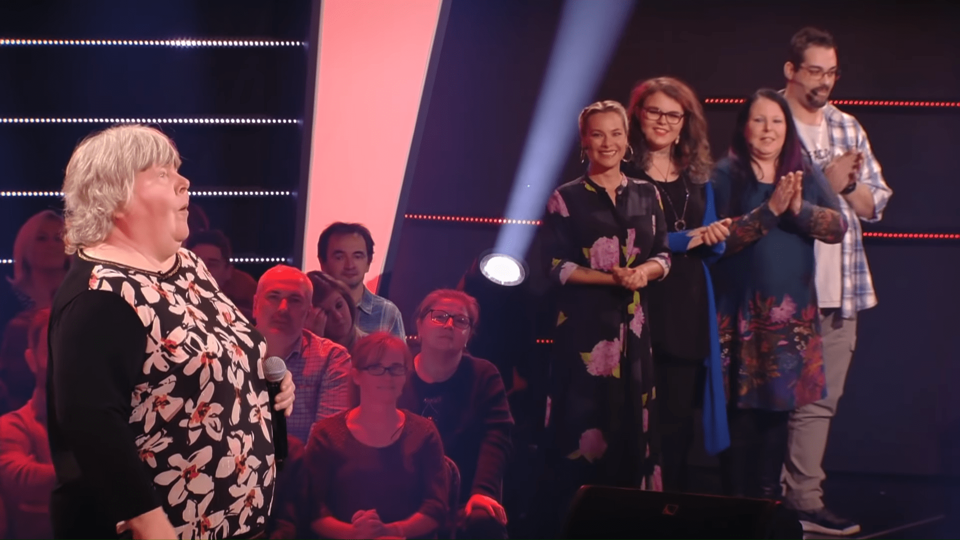 An older adult woman speaks into a microphone in a dimly lit stage setting with an audience clapping. She wears a black and white floral-patterned top.