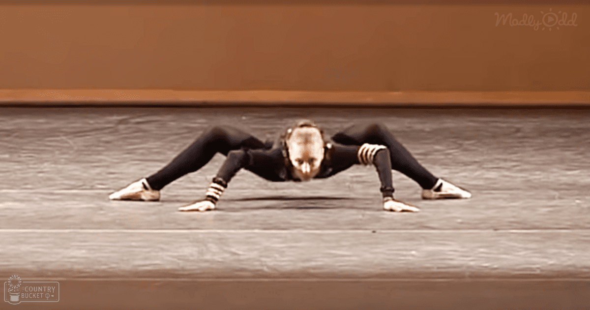 Young adult female wearing a form-fitting black leotard with white stripes, practicing ballet in an indoor dance studio with plain walls.