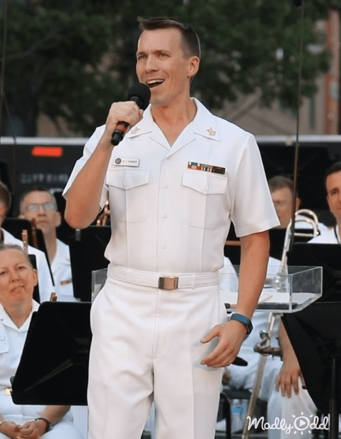 An adult male in a white military uniform speaks into a microphone at an outdoor event with greenery and musical instruments, suggesting a daytime summer or spring setting.