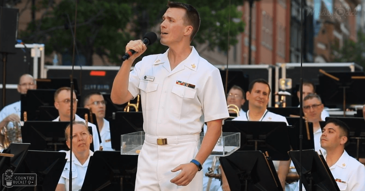 An adult male singer in a white naval uniform performs at an outdoor event with musicians seated on music stands, surrounded by trees and late afternoon light.
