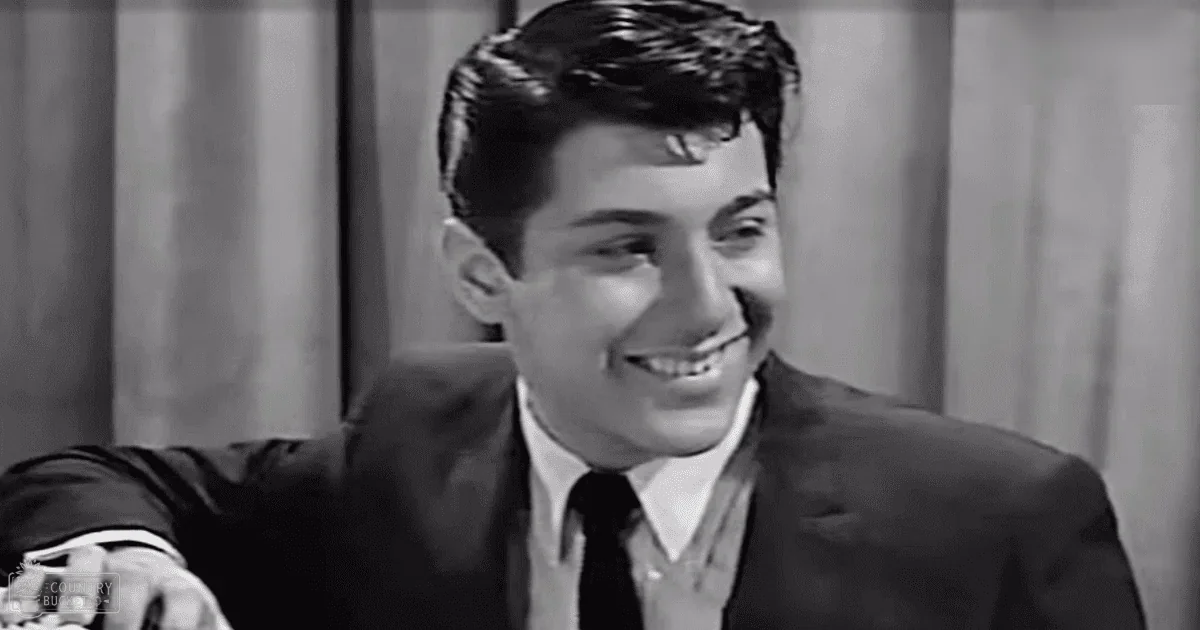 A young adult male wearing a tailored suit jacket and shirt indoors with curtains as part of the background decor.