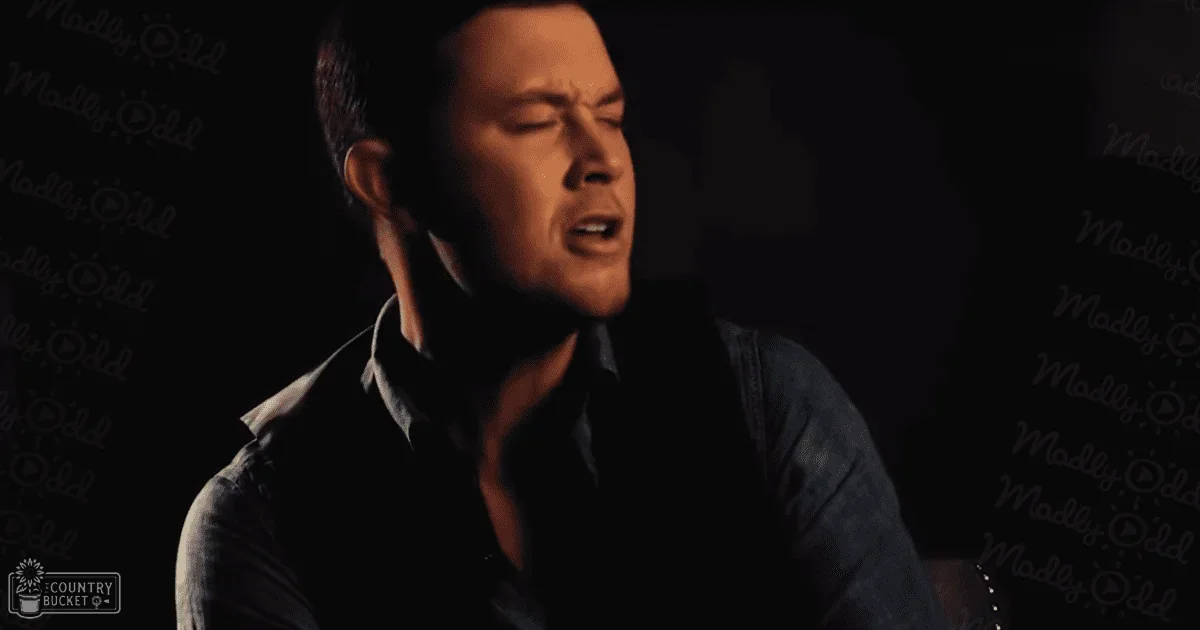 A young adult male sings into a microphone against a dark backdrop with 'Madly Odd' text, suggesting a performance or recording setup.